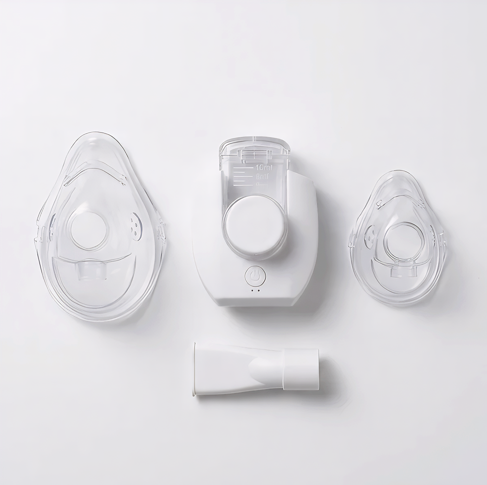A medical breathing device set with a clear face mask, a white central chamber unit, a second small clear mask, and a white connector tube, arranged on a clean white background.