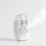 A white and clear handheld nebuliser shown on a clean light gray studio background, with a visible stream of mist coming from the front.
