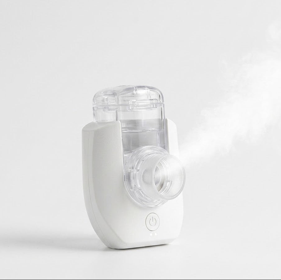 A white and clear handheld nebuliser shown on a clean light gray studio background, with a visible stream of mist coming from the front.