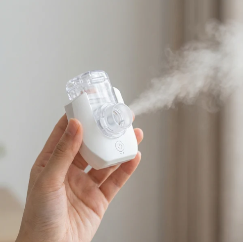 A white and clear handheld nebuliser shown emitting mist against a neutral background